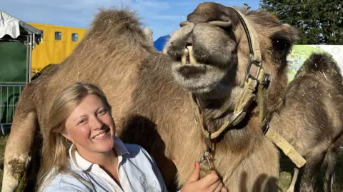 Phil Harrison/BBC A blonde woman smiling and standing next to a large camel.
