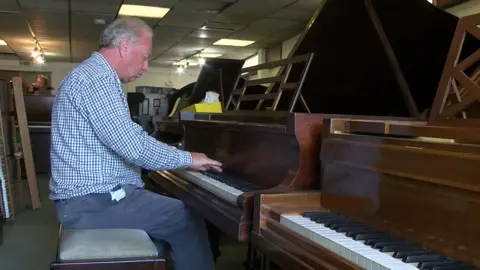 David has a blue-checked collared shirt and blue trousers. He has short grey hair. He is playing a wooden grand piano in the middle of a row of pianos.