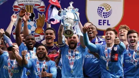 Coventry City's captain Matt Grimes and his team mates lift the Sky Bet Championship trophy during the trophy presentation following the Sky Bet Championship match at the Coventry Building Society Arena, Coventry. Ten players can be seen cheering as the captain, centre, raises the cup, with another holding a bottle of champagne.