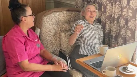 BBC A member of care home staff in a pink tabard sits next to an elderly lady who is sat at a table with a laptop and cup of tea as well as a plate of biscuits.