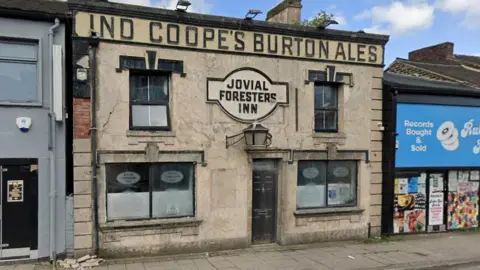 The front of a high street pub. It is a two-storey building with four windows and a door. A light hangs above the door underneath a white sign that says "Jovial Foresters Inn".