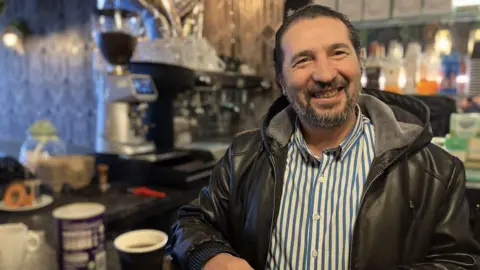 A man with dark hair and a grey beard wearing a leather jacket and a blue and white striped shirt is smiling. He is standing in front of a coffee machine with a cup of coffee beside him. 
