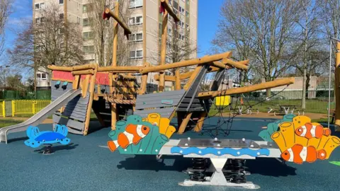 Stevenage Borough Council A play area in front of a block of flats. There is a blue floor, trees in the background and a wooden pirate ship. There is play equipment in front of the ship, with orange and white fish, painted on the front. A blue crab also features to the left on equipment. 