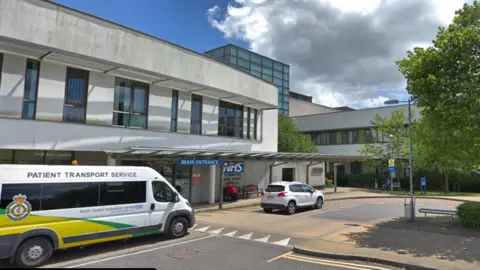 Google An ambulance and a car are parked outside the main entrance to Lymington New Forest Hospital - a grey, two-storey building with a large blue NHS sign on the front