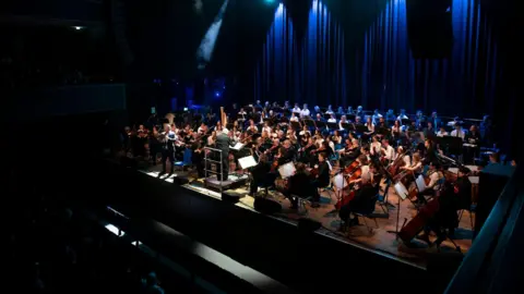 A large orchestra performing on a stage in front of an audience. Blue curtains can be seen behind the stage. The stage is bathed in light while audience members sit in the dark.