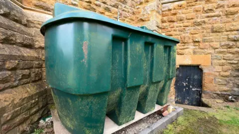 A large green plastic oil tanker stands against a yellow bricked cobbled wall.