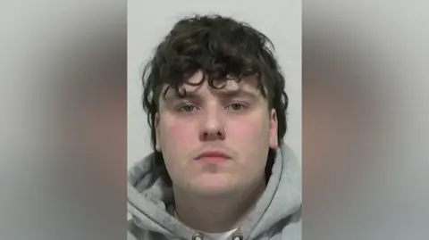 Mugshot of Frier. He is clean shaven with long straggly dark hair. He has a nose ring in his left nostril and is wearing a grey hoody.