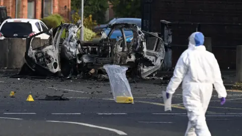 A forensic investigator dressed in a white hazmat suit and purple plastic gloves gathers evidence at the scene of a car bomb attack on Dunmurry Police Station. The investigator is walking towards the wreckage of the vehicle which has been blown apart. Three yellow evidence markers have been placed on the road, one is inside a clear plastic bag. 