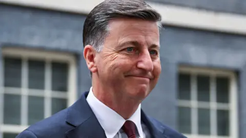 Douglas Alexander wearing a dark blue suit, white shirt and dark red tie walks out of 10 Downing Street.