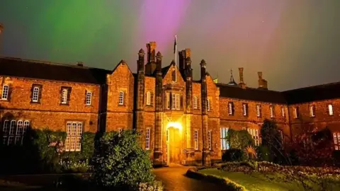 The sky above an old brick building is lit up in blue and green at night time. 