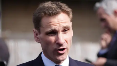 PA Media Chris Philp - a man with short brown hair and wearing a suit - has his mouth partially open while speaking to someone.