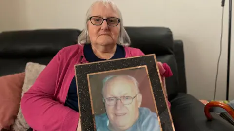 A woman, aged 75, with shoulder-length white hair and black rimmed glasses sits on a sofa looking at the camera. She is holding a framed photograph of her late husband. She is wearing a black top and a raspberry pink cardigan