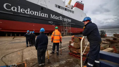 Workers make final preparations ahead of the launch of the MV Glen Rosa in April 2024. Six workers can be seen in the foreground with their back to the camera. The white, black and red ferry, with Caledonian MacBrayne written on the side, is in the distance.
