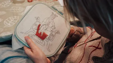 Jersey Heritage A close up of a person embroidering. They are using read thread for an image of a medieval woman holding a child over a cot. 