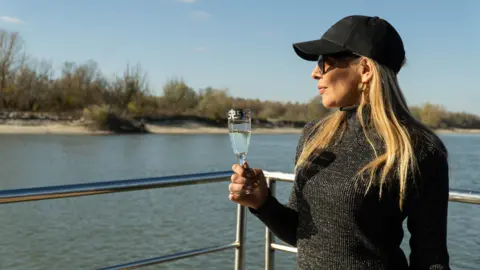 A woman with long blonde hair, wearing a grey jumper and a dark baseball hat, drinks champagne from a fluted glass while looking out at a desolate landscape next to a river
