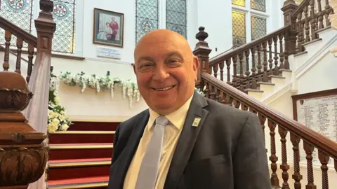 Andrew Turner/BBC Council leader Carl Smith, standing on the staircase at Great Yarmouth He is wearing a dark grey suit, light grey tie, and white shirt. He is bald, and smiling. 