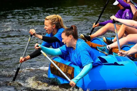 John  Mcintyre Two woman, one with long blonde hair and one with long brown hair, both in ponytails, are leaning out in front of a boat pulling it along with oars.