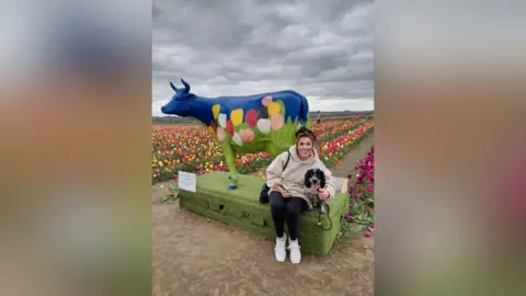 Jemma Ladwitch A woman wearing black leggings, white trainers and a pale pink hoodie posing next to a statue of a blue cow with a tulip field painted on it with her arm around a black and white spaniel.