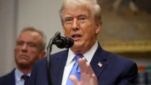 Getty Images US President Donald Trump (R) talking in the White House on Monday 22 September 2025, with US secretary of Health and Human Services Robert F. Kennedy Jr (L) in the background. Both men wear dark suits and ties. 