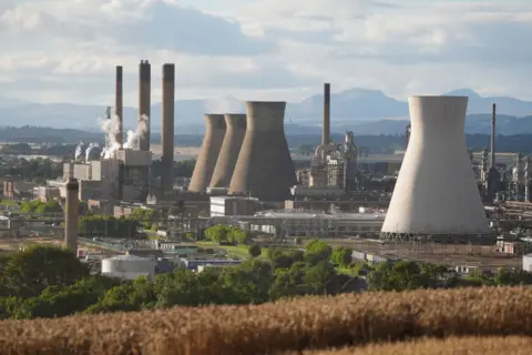 PA Media A view of the Grangemouth oil refinery with several large chimneys visible and smaller industrial buildings sitting beneath them. The complex is sitting against the backdrop of trees and hills in the distance. 