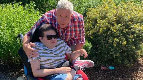 A man with white hair stands next to a younger man sitting in a wheelchair wearing sunglasses.