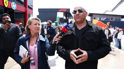 Getty Images Vin Diesel being interviewed. He is wearing a black jacket and black top and black sunglass with a smile on his face. The female interviewer is wearing a pink top and blue coat. An onlooker takes a picture on their mobile phone. They are all in a busy paddock area.
