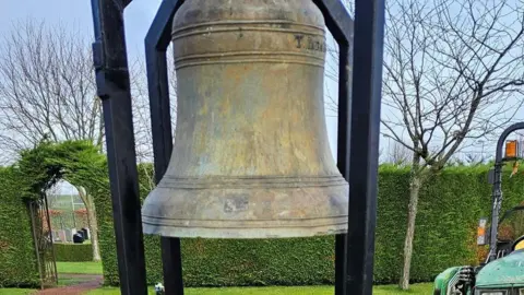 A weathered bronze bell is being placed in a cemetery and a green tractor is in the background.