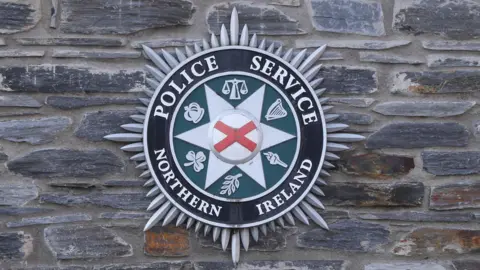 The Police Service of Northern Ireland badge, an emblem with white, red, green and black colouring that is mounted on a stone wall.
