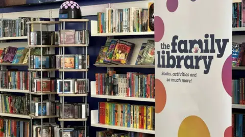 A tall banner reads "The Family Library: books, activities and so much more!" It is white with multicoloured spots and is standing in front of a bookshelf full of books.