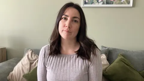 A woman with dark, shoulder-length hair sat on a sofa in her living room wearing a grey long sleeved T-shirt. 