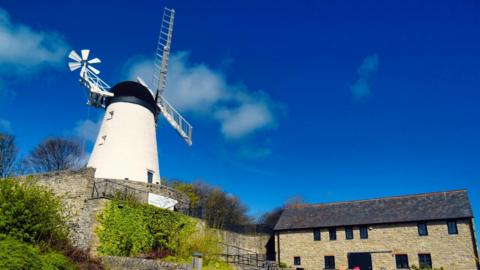 A general view of Fulwell Windmill, with a two-stoey building to the right and a blue sky dotted with clouds above