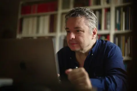 BBC/Katie Razzall Dr Gradel looking at his laptop. 
Wearing dark blue shirt
Bookcase behind him.