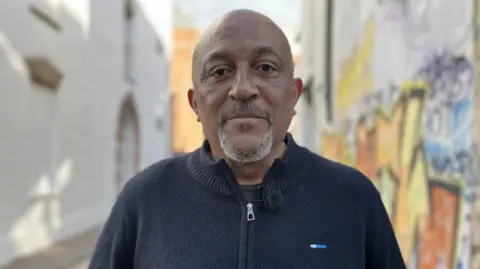 Desmond Brown with blue zip-up sweater. Bald, middle-aged, with a white beard. Behind it is a street with lots of colorful graffiti on it. 