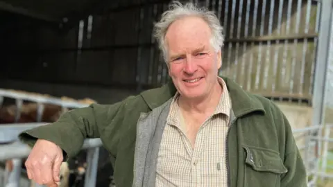 A man with white hair smiles at the camera. He is leaning on a metal farm gate. He is wearing a brown, plaid shirt and dark green jacket. In the background is an open-sided barn.