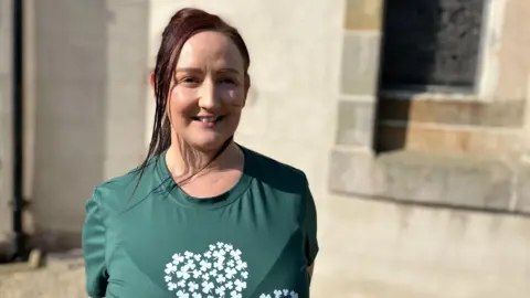 Paula Reid is wearing a green tee-shirt with a shamrock on it. She is smiling directly at the camera and standing in front of the Ramoan Church of Ireland building in Ballycastle. Paula has dark hair which is tied back.