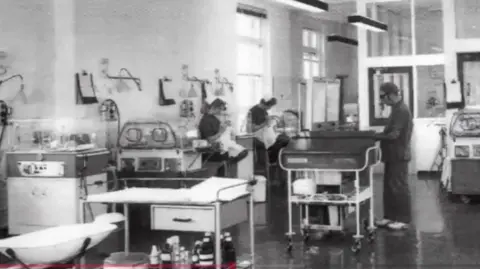 Alder Hey A black-and-white photograph of a hospital ward taken at the old building on Myrtle Street in Liverpool. A number of medics are standing over incubators and are surrounded by medical equipment. 