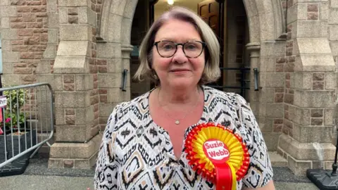 A white women with blonde hair in a bob hairstyle. She has black glasses with big rims. She's wearing a white top with gold and black patterns, and wears a large red and yellow rosette on the left of her shirt which says her name: 'Suzie Webb'. She's standing in front of a large, arched brick building entrance.