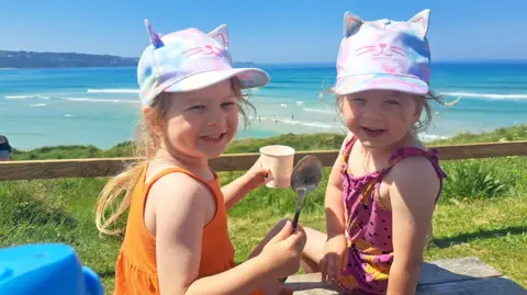 Brioney Garrett Identical twins Nancy and Margo eat ice cream at the beach, wearing colourful caps and strappy tops, with azure-blue sea and gentle waves visible in the background.