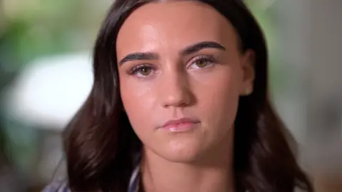 A young woman with long brown hair looks into the camera with a serious expression