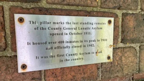 Metal plaque explaining that the brick pillar it is mounted on is the last fragment of the original Nottingham Asylum
