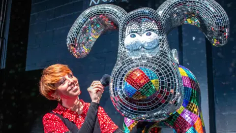 A mosaic Gromit sculpture made out of clear and coloured glass tiles. The eyes are looking up and a woman with short ginger hair and wearing a red polka dot top is smiling whilst brushing the sculpture.