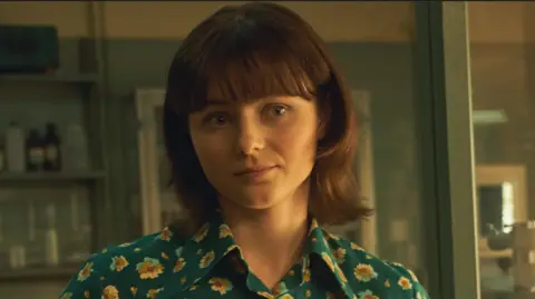 Netflix Thomasin McKenzie looking at the camera. She has brown hair and blue shirt with sunflowers on it. She is stood in a laboratory.