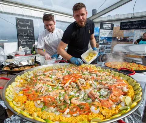 Pete Robinson A man with short hair and a beard stands behind a giant plate of paella