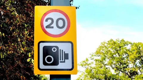 A road sign showing a 20mph speed limit symbol above a speed camera symbol on a yellow background. In the background are trees and a section of blue sky. 