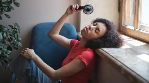 Woman in red top sits against a wall with a handheld fan. She has her eyes closed and her head rested on a windowsill. 