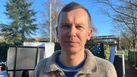 BBC Michael Bradley, dressed in a casual jumper, looking at the camera, with piles of paperwork and school equipment behind.