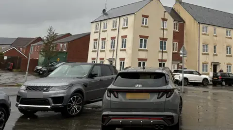Two silver cars parked next to each other on a pavement in a residential area. There's a mixture of red brick and white houses. Other cars are parked on the side of the road. 