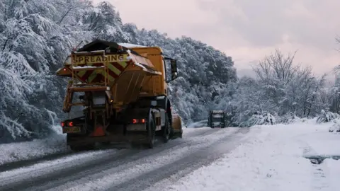 BBC Weather Watchers/Ewan1990 Two gritting trucks treat a snowy road surrounded by evergreen trees. The sky is cloudy.