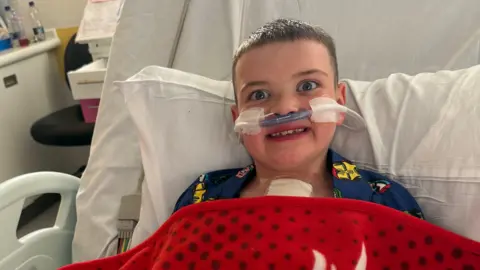 A young boy is pictured in a hospital bed. He is dressed in pyjamas and has a red patterned blanket on top. He has 