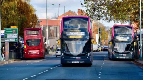 Three buses travelling on a major Birmingham road, with a signpost for city centre, City Hospital and Handsworth visible on the left. The double-decker buses approaching the camera are the Bearwood number 82 and Dudley number 87 services.